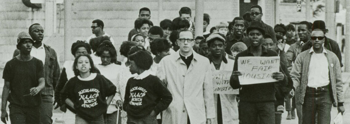 A group of predominantly young, African-American protesters with a white priest in the center of the group.